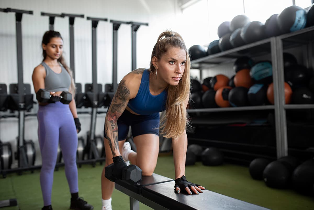 Dos mujeres entrenando con pesas en un gimnasio; una realiza remo con mancuerna sobre un banco y la otra usa mancuernas de pie